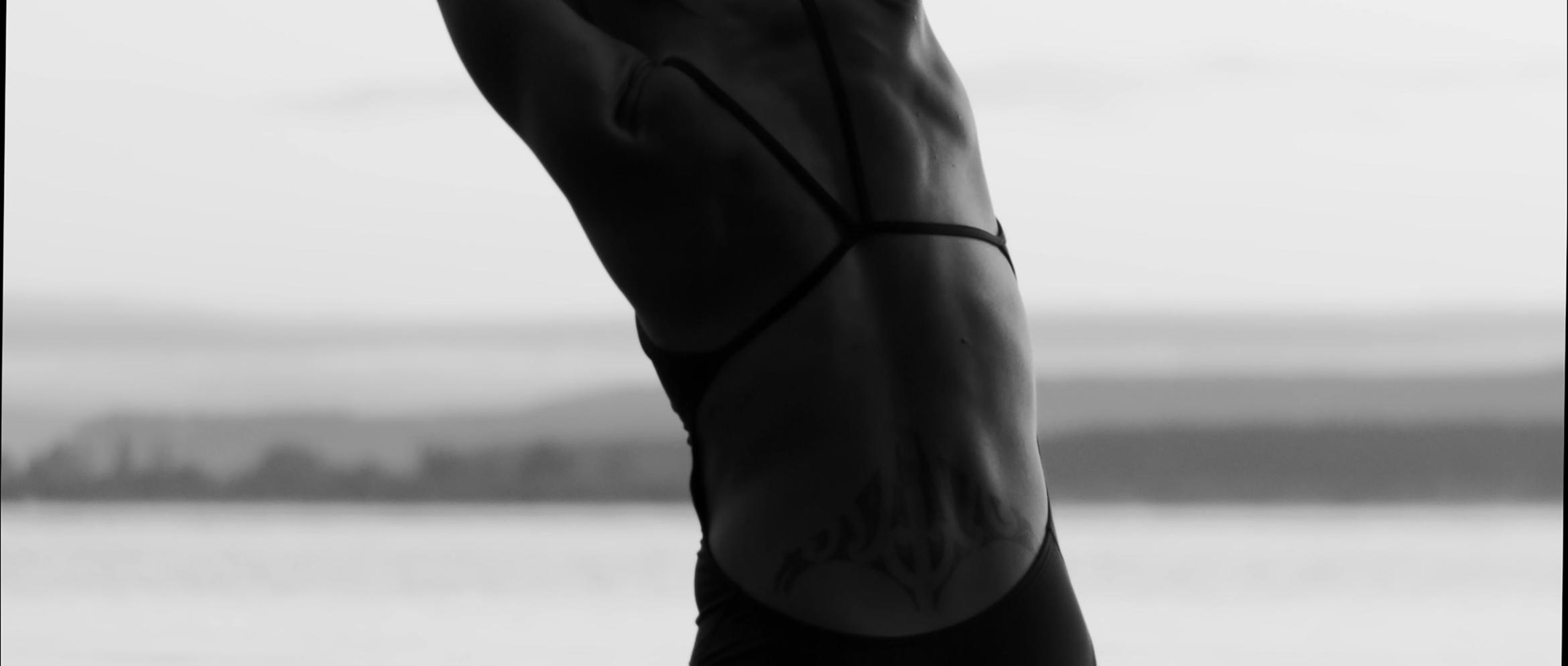 The back of a women swimmer, wearing black togs and stretching to the left.